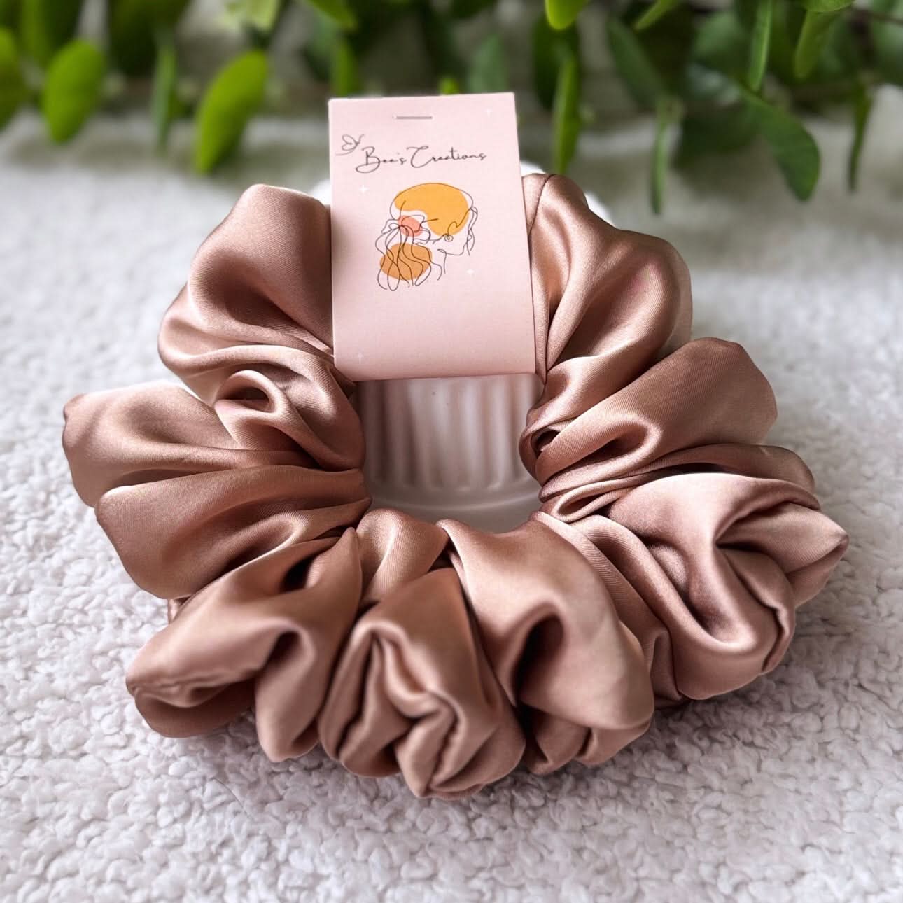 Pink satin scrunchie with a branded card on a textured surface with greenery in the background