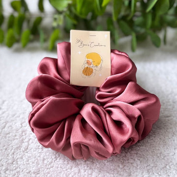 Pink scrunchie with a branded card on a textured surface with greenery in the background