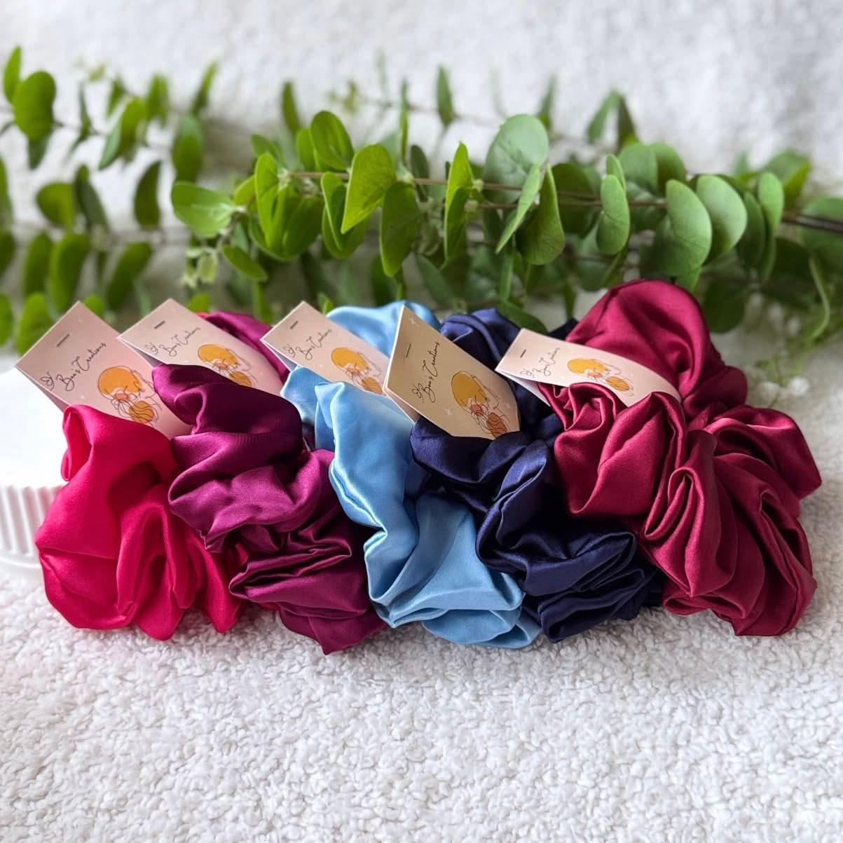 Four colorful scrunchies on a white surface with green leaves in the background
