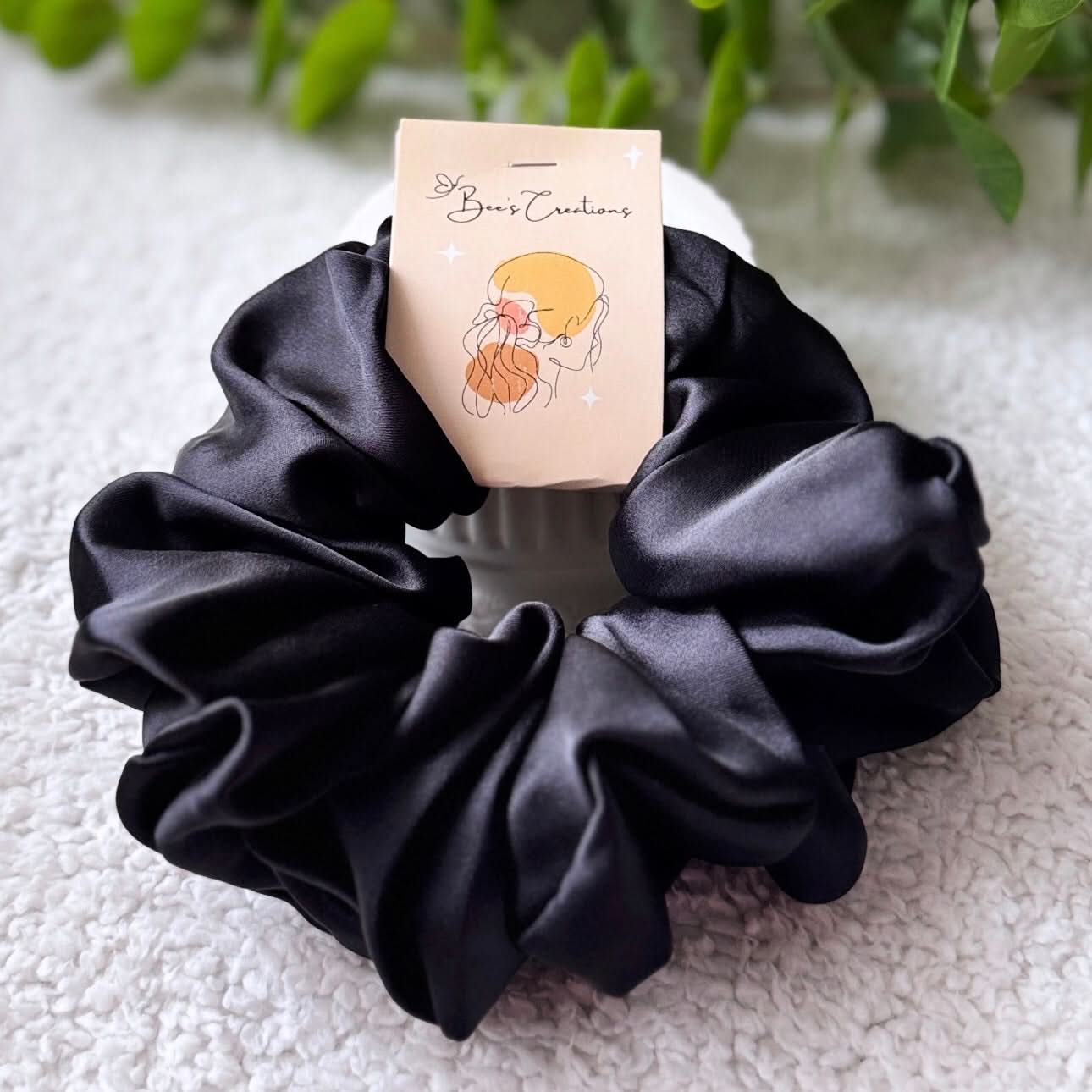 Black satin scrunchie with a branded card on a textured surface with greenery in the background