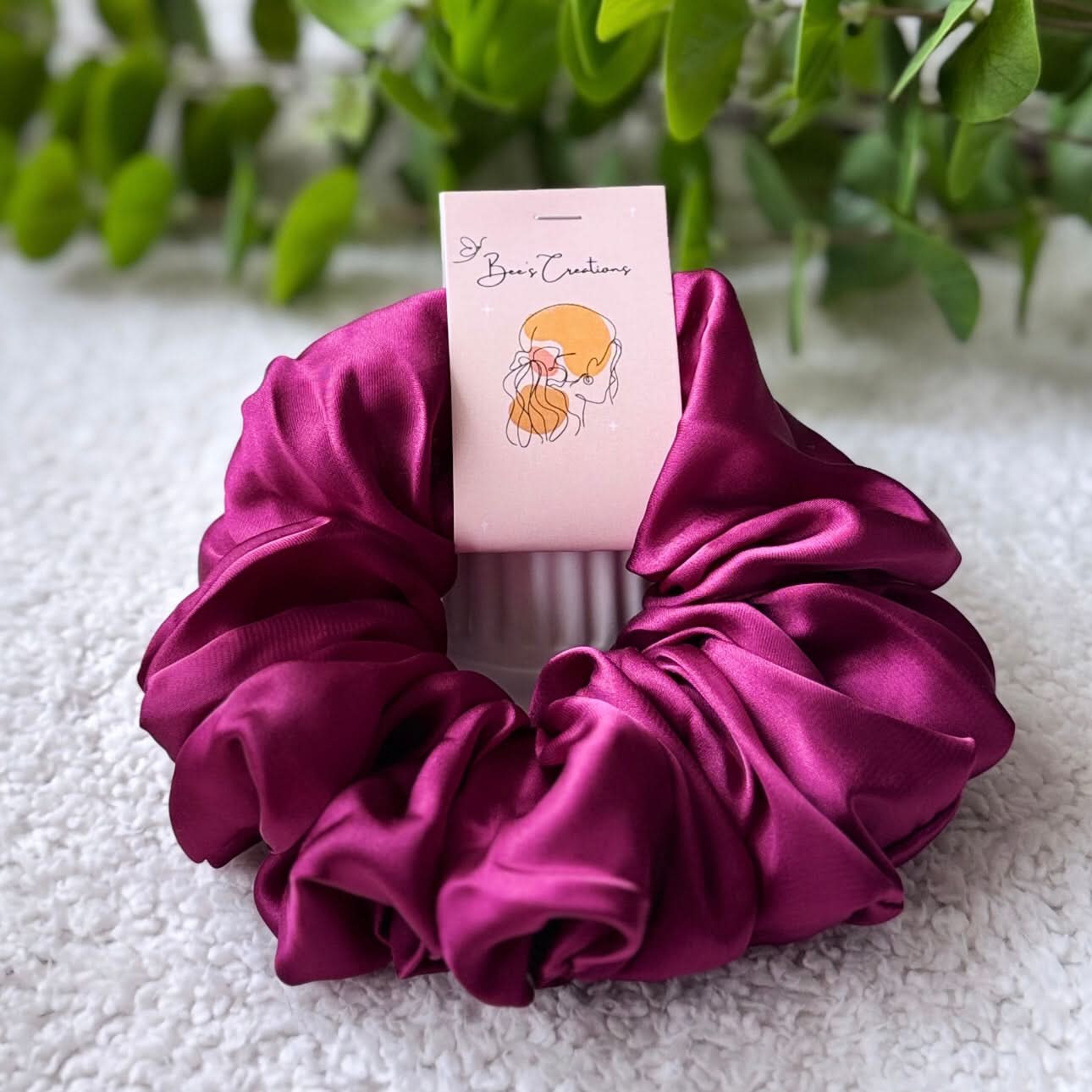 Purple satin scrunchie with a pink card on a textured surface with greenery in the background
