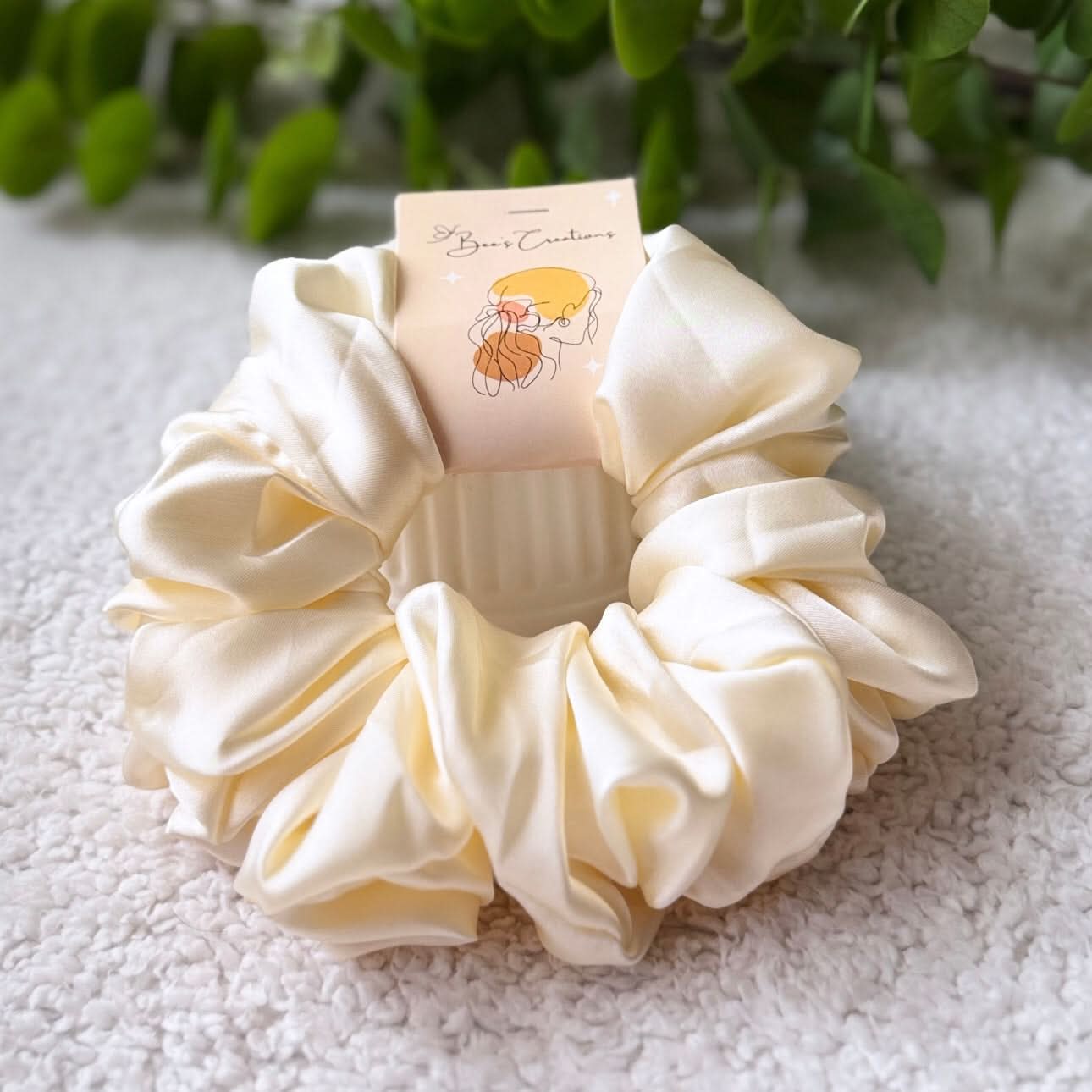 Cream-colored scrunchie with a small card on a textured surface with greenery in the background