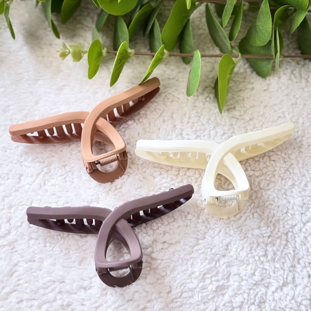 Three hair clips in beige, brown, and white on a textured surface with green leaves in the background.