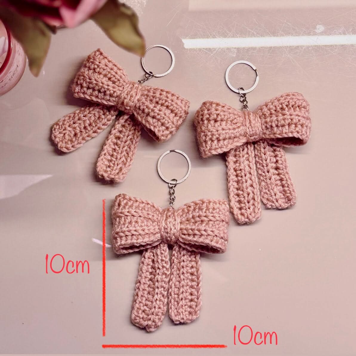 Three pink crocheted bow keychains on a light pink background with measurements indicated.