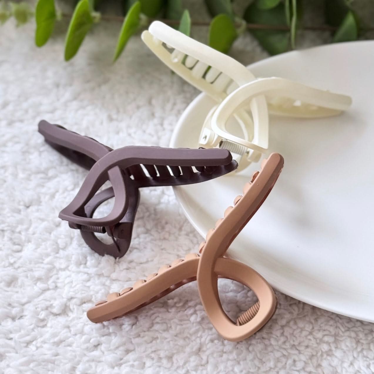Three hair clips in brown, beige, and rose gold on a textured surface with green leaves in the background.