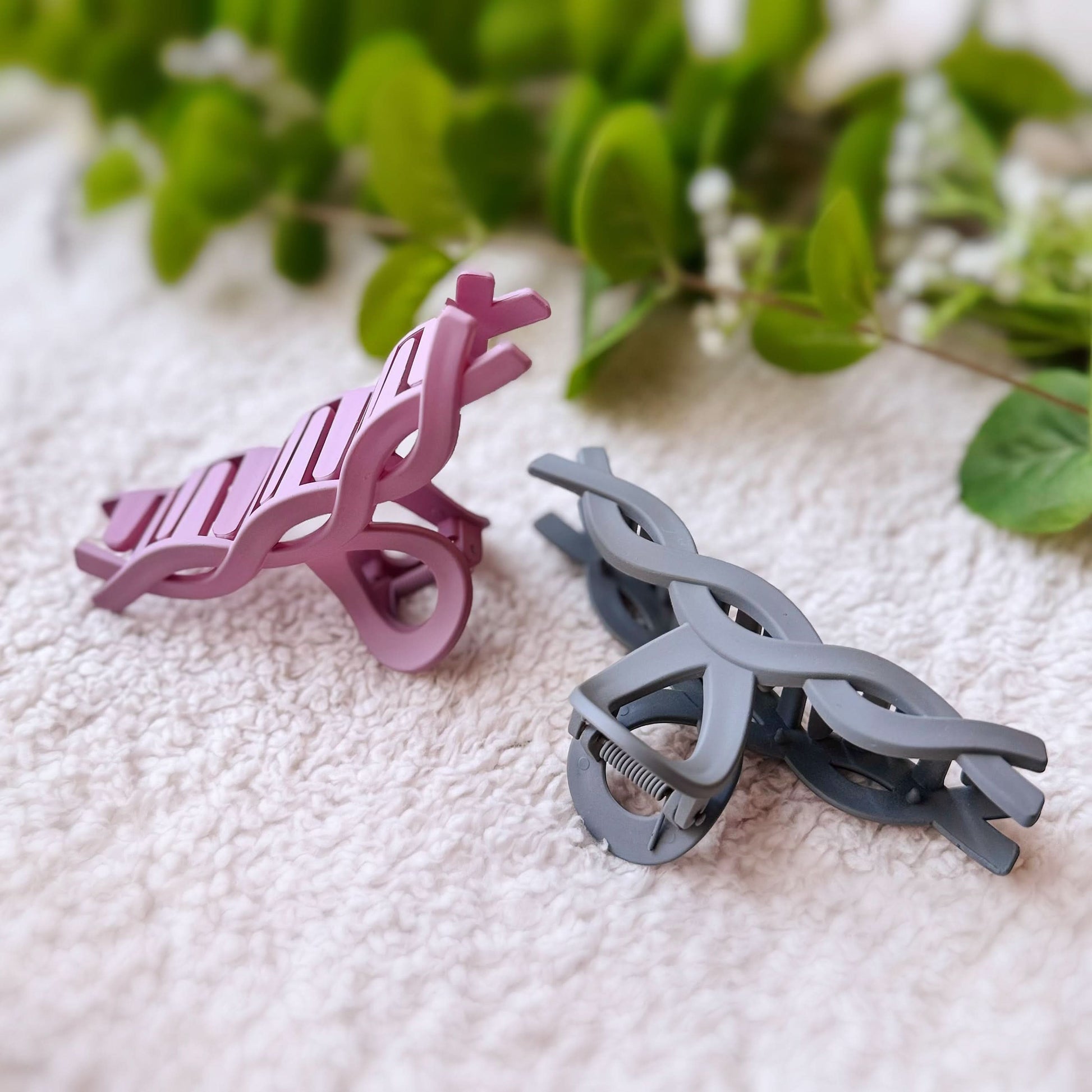 Two hair clips, one purple and one gray, on a textured surface with green leaves in the background.
